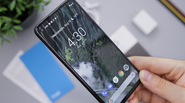 En hand håller upp en Android-mobil över ett skrivbord, med hemskärmen aktiv.
