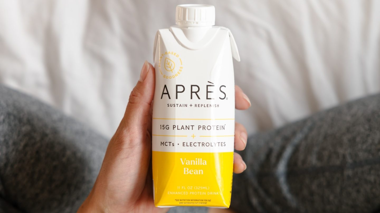 En hand håller upp en färdig proteinshake över ett sänglakan.