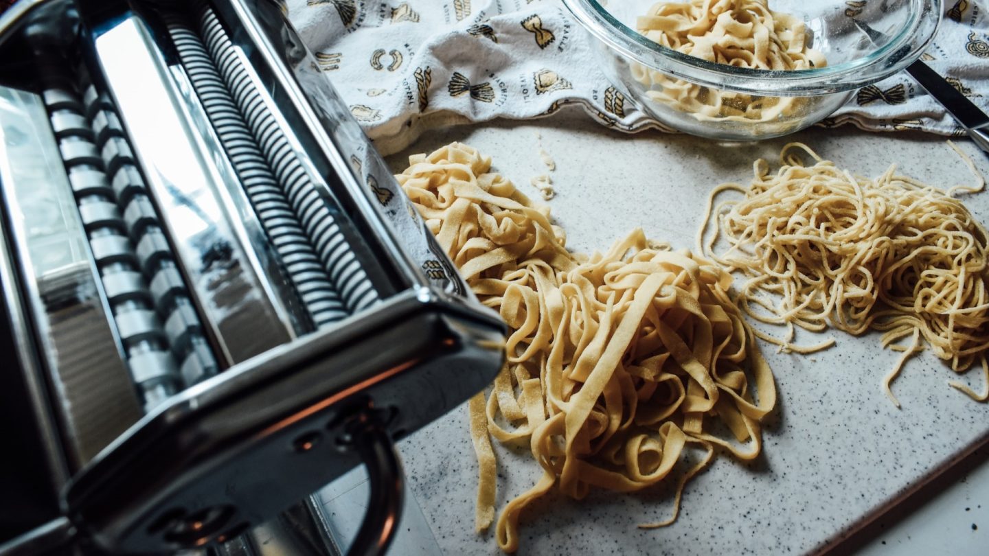 En pastamaskin står på en köksbänk bredvid några högar med färdig pasta.