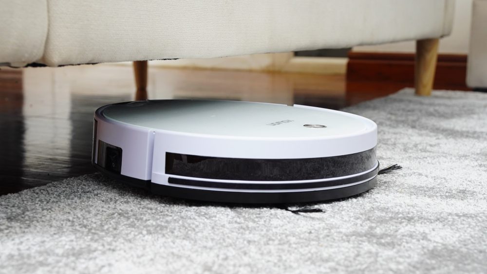 En vit robotdammsugare håller på att dammsuga en matta vid kanten av en soffa.
