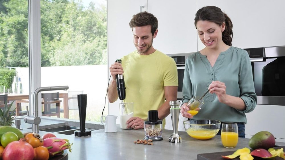 En man och en kvinna står bredvid varandra i ett kök och gör smoothies med en stavmixer.